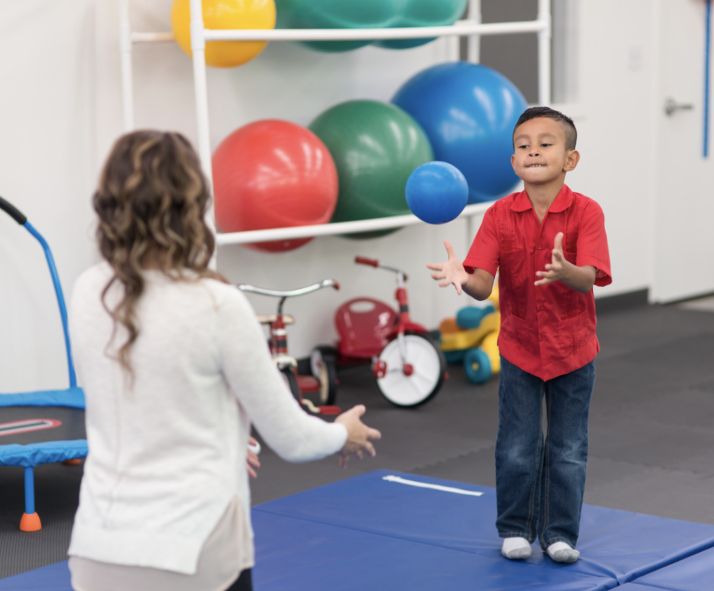 chłopiec siedzi na piłce fitnessowej podtrzymywany przez terapeutkę
